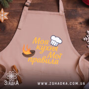 Фартух моя кухня, бежевий колір, зображення з верхнього ракурсу, фон дерев'яний, принт з написом 'Моя кухня Мої правила' та зображенням шеф-капелюха і вогню.
