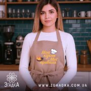 Фартух моя кухня, бежевий колір, зображення з фронтального ракурсу, фон сучасної кухні з темно-синьою плиткою, принт з написом 'Моя кухня Мої правила' та зображенням шеф-капелюха і вогню.