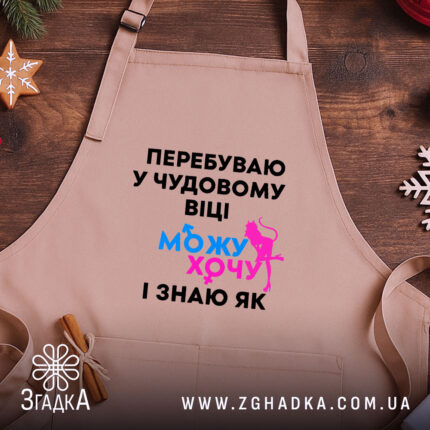 Фартух можу і хочу, бежевого кольору, зображений на дерев'яному фоні, деталі принта чорного кольору з яскравими елементами синього та рожевого.
