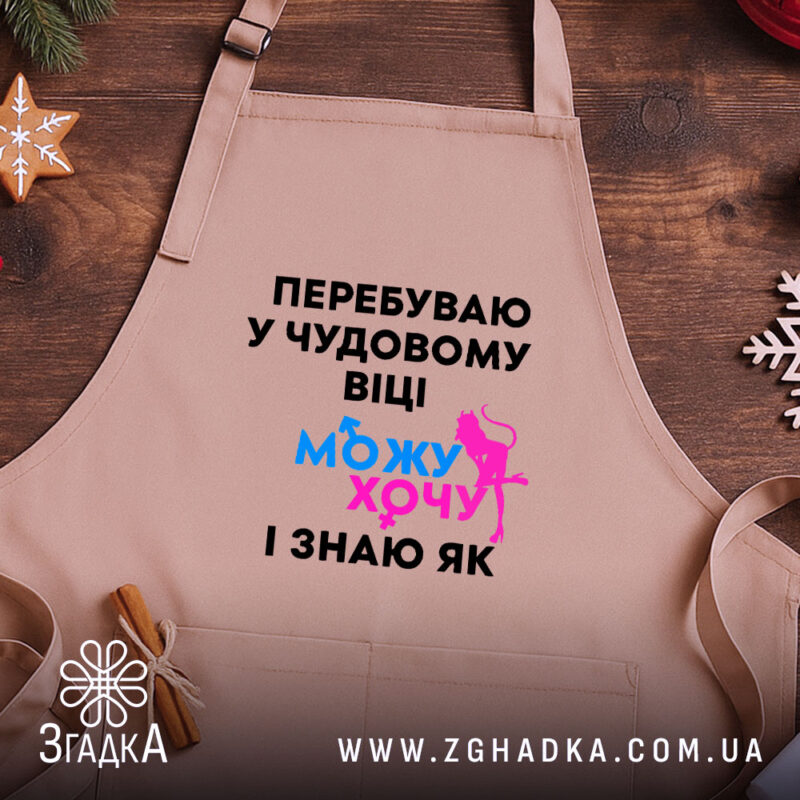 Фартух можу і хочу, бежевого кольору, зображений на дерев'яному фоні, деталі принта чорного кольору з яскравими елементами синього та рожевого.