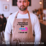 Фартух можу і хочу, бежевого кольору, зображений на чоловікові в кафе, фон розмитий, принт чорного кольору з яскравими елементами синього та рожевого.