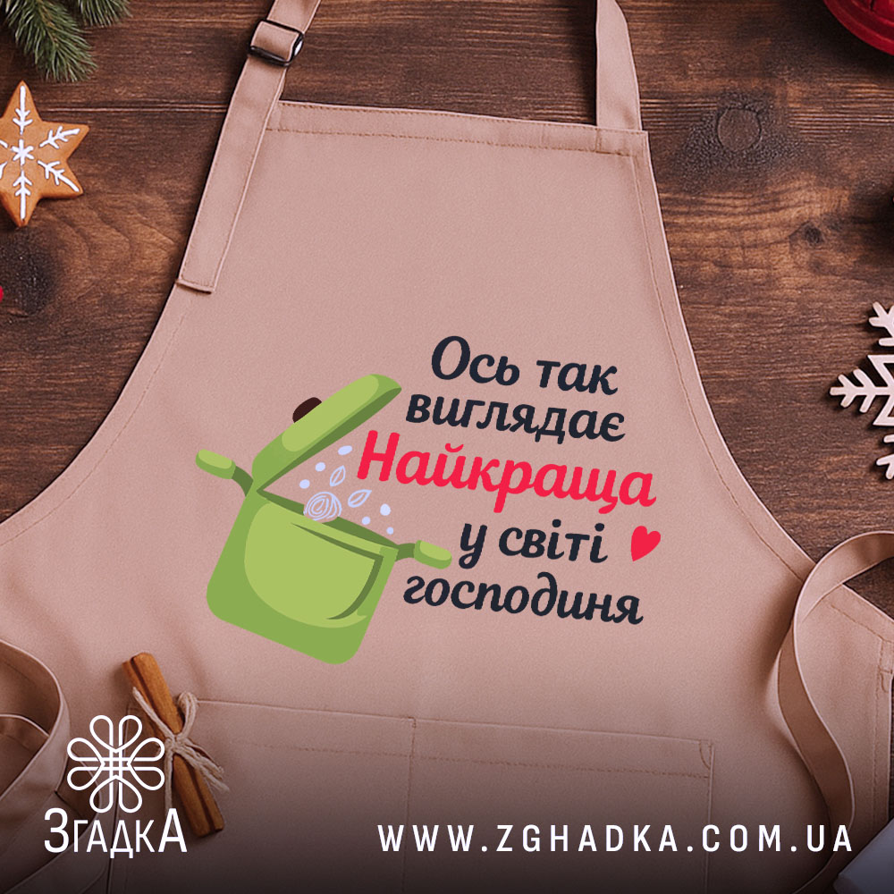 Фартух найкраща господиня, бежевого кольору, зображений з близького ракурсу на дерев'яному фоні, принт з написом "Ось так виглядає Найкраща у світі господиня" та малюнком каструлі, навколо декор у вигляді новорічних зірочок.