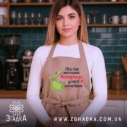 Фартух найкраща господиня, бежевого кольору, зображений на жінці з довгим волоссям, фон кухонний з темно-синьою стіною, принт з написом "Ось так виглядає Найкраща у світі господиня" та малюнком каструлі.