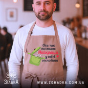 Фартух найкраща господиня, бежевого кольору, зображений на чоловікові з коротким волоссям, фон кафе з розмитими силуетами людей, принт з написом "Ось так виглядає Найкраща у світі господиня" та малюнком каструлі.