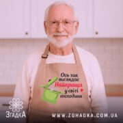 Фартух найкраща господиня, бежевого кольору, зображений з близького ракурсу на дерев'яному фоні, принт з написом "Ось так виглядає Найкраща у світі господиня" та малюнком каструлі, навколо декор у вигляді новорічних зірочок.