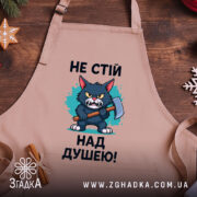 Фартух не стій над душею, бежевого кольору, зображений з близького ракурсу на дерев'яному фоні, принт з малюнком кота з сокирою та текстом, навколо новорічні елементи декору.
