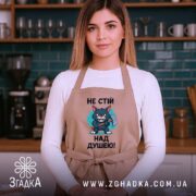 Фартух не стій над душею, бежевого кольору, зображений на жінці в кухонному інтер'єрі, фон з дерев'яними полицями та кухонним приладдям, принт з малюнком кота з сокирою та текстом.