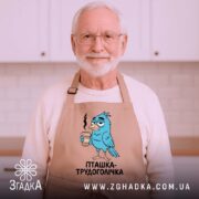 Фартух пташка трудоголічка, бежевого кольору, зображений на дерев'яному фоні, принт з пташкою, що тримає каву, з димком над чашкою, декор у вигляді новорічних елементів.