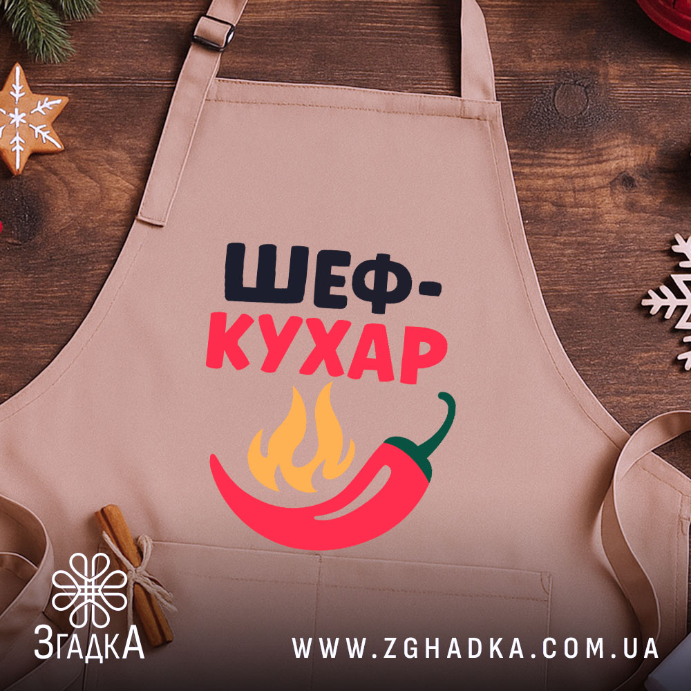 Фартух шеф кухар бежевого кольору, зображений зверху на дерев'яному фоні, принт з написом 'ШЕФ-КУХАР' та червоним перцем і вогнем, поруч з декором у вигляді новорічних зірочок.
