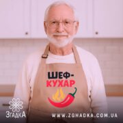 Фартух шеф кухар бежевого кольору, зображений зверху на дерев'яному фоні, принт з написом 'ШЕФ-КУХАР' та червоним перцем і вогнем, поруч з декором у вигляді новорічних зірочок.
