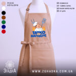 Фартух татко всіх нагодує. Фартух бежевого кольору, зображений з фронтального ракурсу на манекені. Фон світлий, без зайвих деталей. Принт містить малюнок кухаря з лопаткою та виделкою, а також напис 'ТАТКО ВСІХ НАГОДУЄ' у яскравих кольорах.
