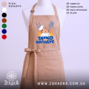 Фартух татко всіх нагодує. Фартух бежевого кольору, зображений з фронтального ракурсу на манекені. Фон світлий, без зайвих деталей. Принт містить малюнок кухаря з лопаткою та виделкою, а також напис 'ТАТКО ВСІХ НАГОДУЄ' у яскравих кольорах.