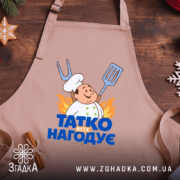 Фартух татко всіх нагодує. Бежевий фартух, зображений з близького ракурсу на дерев'яному фоні. На фоні видно декор у вигляді новорічних зірочок. Принт з кухарем та написом 'ТАТКО ВСІХ НАГОДУЄ' яскравий і помітний.