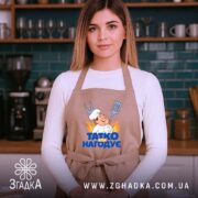 Фартух татко всіх нагодує. Бежевий фартух, зображений на жінці, яка стоїть у кухні. Фон складається з дерев'яних поличок з кухонним приладдям. Принт з кухарем та написом 'ТАТКО ВСІХ НАГОДУЄ' добре видимий.