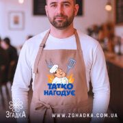 Фартух татко всіх нагодує. Бежевий фартух, зображений на чоловікові, який стоїть у кафе. Фон розмитий, з видимими елементами інтер'єру. Принт з кухарем та написом 'ТАТКО ВСІХ НАГОДУЄ' яскравий і привертає увагу.