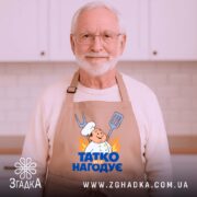 Фартух татко всіх нагодує. Бежевий фартух, зображений з близького ракурсу на дерев'яному фоні. На фоні видно декор у вигляді новорічних зірочок. Принт з кухарем та написом 'ТАТКО ВСІХ НАГОДУЄ' яскравий і помітний.