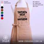 Фартух татусь усіх нагодує, бежевого кольору, зображений з фронтального ракурсу на манекені, фон світлий, принт чорного кольору з написом та малюнком вусів.