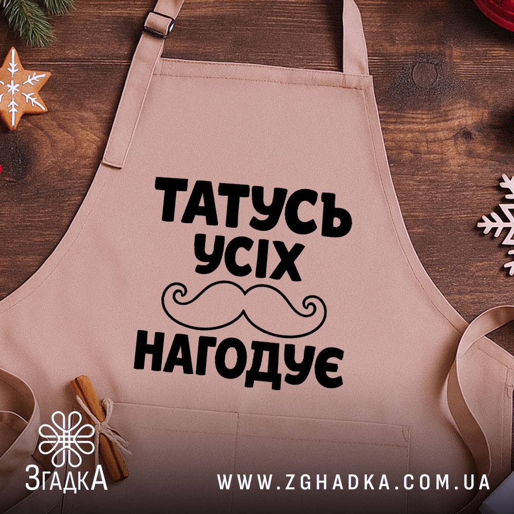 Фартух татусь усіх нагодує, бежевого кольору, зображений з верхнього ракурсу на дерев'яному фоні, деталі принта чорного кольору з написом та вусами, навколо декор у вигляді новорічних зірочок.