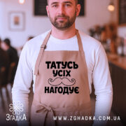 Фартух татусь усіх нагодує, бежевого кольору, зображений на чоловікові в кафе, фон розмитий, принт чорного кольору з написом та вусами, чоловік дивиться в камеру.