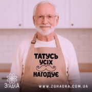 Фартух татусь усіх нагодує, бежевого кольору, зображений з верхнього ракурсу на дерев'яному фоні, деталі принта чорного кольору з написом та вусами, навколо декор у вигляді новорічних зірочок.