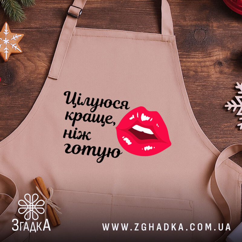Фартух цілуюся краще ніж готую, бежевого кольору, зображений з верхнього ракурсу на дерев'яному фоні, деталі принта включають червоні губи та чорний текст, поряд з декором у вигляді зірочки та кориці.