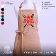 Фартух жарю як треба, бежевого кольору, зображений на манекені з ракурсу спереду, фон світлий, принт з червоним текстом та графічними елементами у вигляді вогню та кухонних інструментів.