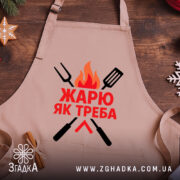 Фартух жарю як треба, бежевого кольору, зображений зверху на дерев'яному фоні, принт з червоним текстом та графічними елементами у вигляді вогню та кухонних інструментів, навколо розташовані святкові елементи.