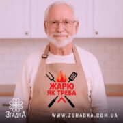 Фартух жарю як треба, бежевого кольору, зображений зверху на дерев'яному фоні, принт з червоним текстом та графічними елементами у вигляді вогню та кухонних інструментів, навколо розташовані святкові елементи.