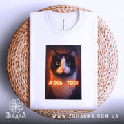 Футболка а ось тобі біла, складена на плетеній кошику, ракурс зверху, принт кота з написом "А ОСЬ ТОБІ" на видному місці.