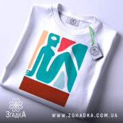 Футболка абстрактне арт зображення, біла, складена на поверхні, фон світло-сірий, принт зображує стилізовану фігуру людини в блакитному, червоному та бежевому кольорах, з етикеткою на шнурку.