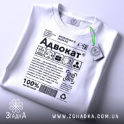 Футболка адвокат білого кольору, зображена зверху на столі, фон світлий, принт з текстом та символами, на комірці є етикетка з зеленим шнурком.