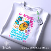 Футболка акушер, біла, зображена на столі, фон світло-сірий, принт з мультяшним малюнком немовляти в ковдрі, з червоним серцем та кольоровими елементами, на комірці є етикетка.