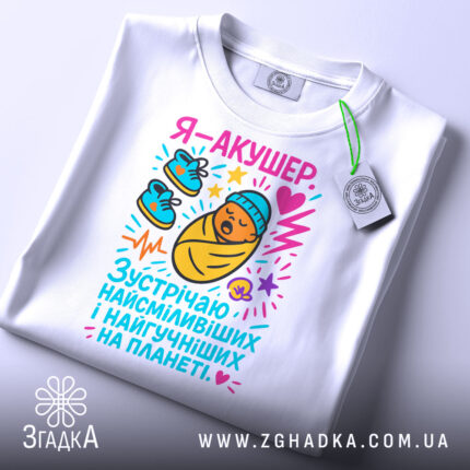 Футболка акушер, біла, зображена на столі, фон світло-сірий, принт з мультяшним малюнком немовляти в ковдрі, з червоним серцем та кольоровими елементами, на комірці є етикетка.