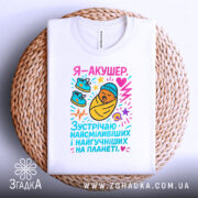 Футболка акушер, біла, зображена на столі, фон світло-сірий, принт з мультяшним малюнком немовляти в ковдрі, з червоним серцем та кольоровими елементами, футболка складена на плетеному підносі.