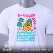 Футболка акушер, біла, зображена на столі, фон світло-сірий, принт з мультяшним малюнком немовляти в ковдрі, з червоним серцем та кольоровими елементами, на комірці є етикетка.