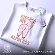 Футболка альпака гумор, біла, складена на столі, фон світло-сірий, принт з альпакою та написом 'KISS MY ALPACASS' у рожевих тонах, з етикеткою зеленого кольору.