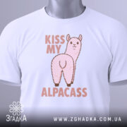 Футболка альпака гумор, біла, складена на столі, фон світло-сірий, принт з альпакою та написом 'KISS MY ALPACASS' у рожевих тонах, з етикеткою зеленого кольору.