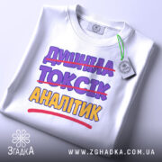 Футболка аналітик не токсік, біла, складена на поверхні, фон світло-сірий, принт з текстом "ДИШИНА ТОКСІК АНАЛІТИК" з перекресленим словом "ТОКСІК", кольори фіолетовий та жовтий, на комірці є етикетка.