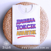 Футболка аналітик не токсік, біла, складена на плетеній кошику, фон світлий, принт містить текст "ДИШИНА ТОКСІК АНАЛІТИК" з перекресленим словом "ТОКСІК", кольори фіолетовий та жовтий.