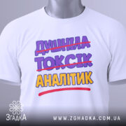 Футболка аналітик не токсік, біла, складена на поверхні, фон світло-сірий, принт з текстом "ДИШИНА ТОКСІК АНАЛІТИК" з перекресленим словом "ТОКСІК", кольори фіолетовий та жовтий, на комірці є етикетка.