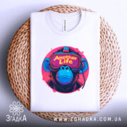 Футболка Another Life білого кольору, складена на плетеній кошику, ракурс зверху, фон світло-сірий, принт з малюнком мавпи в окулярах віртуальної реальності, яскраві кольори принта: рожевий, синій, жовтий.