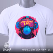 Футболка Another Life білого кольору, складена на поверхні, ракурс зверху, фон світло-сірий, принт з малюнком мавпи в окулярах віртуальної реальності, яскраві кольори принта: рожевий, синій, жовтий, на етикетці зелена стрічка.