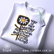 Футболка антивоєнна позиція, біла, складена на столі, фон світло-сірий, принт з жовтою квіткою та текстом чорного кольору, з етикеткою зеленого кольору.