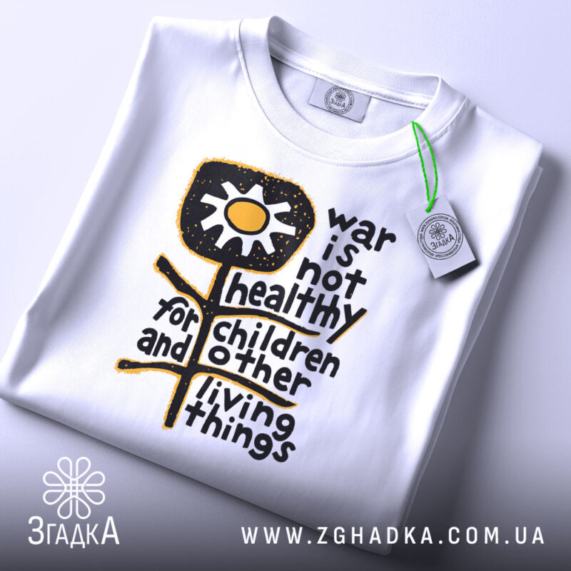Футболка антивоєнна позиція, біла, складена на столі, фон світло-сірий, принт з жовтою квіткою та текстом чорного кольору, з етикеткою зеленого кольору.