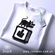 Футболка антиЗМІ білого кольору, складена на поверхні, з принтом чорного кольору, що зображає мишу на телевізорі з жестом середнього пальця, на футболці є етикетка з зеленим шнурком, фон світло-сірий.