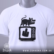 Футболка антиЗМІ білого кольору, складена на поверхні, з принтом чорного кольору, що зображає мишу на телевізорі з жестом середнього пальця, на футболці є етикетка з зеленим шнурком, фон світло-сірий.