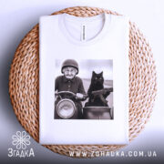 Футболка бабуся і автомат. Біла футболка, складена на плетеній основі, з принтом бабусі на мотоциклі з котом. Фон світлий, принт контрастний, з деталями обличчя та одягу бабусі.