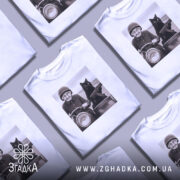 Футболка бабуся і автомат. Біла футболка з принтом, на якому зображена бабуся на мотоциклі з чорним котом, що тримає автомат. Фон розмитий, з легкими відтінками сірого. Принт чіткий, з деталями обличчя бабусі та кота.