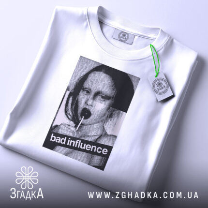 Футболка bad influence білого кольору, складена на поверхні, фон світлий, принт з дівчиною, що тримає льодяник, з текстом 'bad influence', на комірці є етикетка.