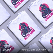 Футболка байкер мама сила. Біла футболка з принтом, на якому зображена жінка на мотоциклі, ракурс спереду, фон світло-сірий, принт виконаний у яскравих кольорах: червоний, чорний, рожевий.